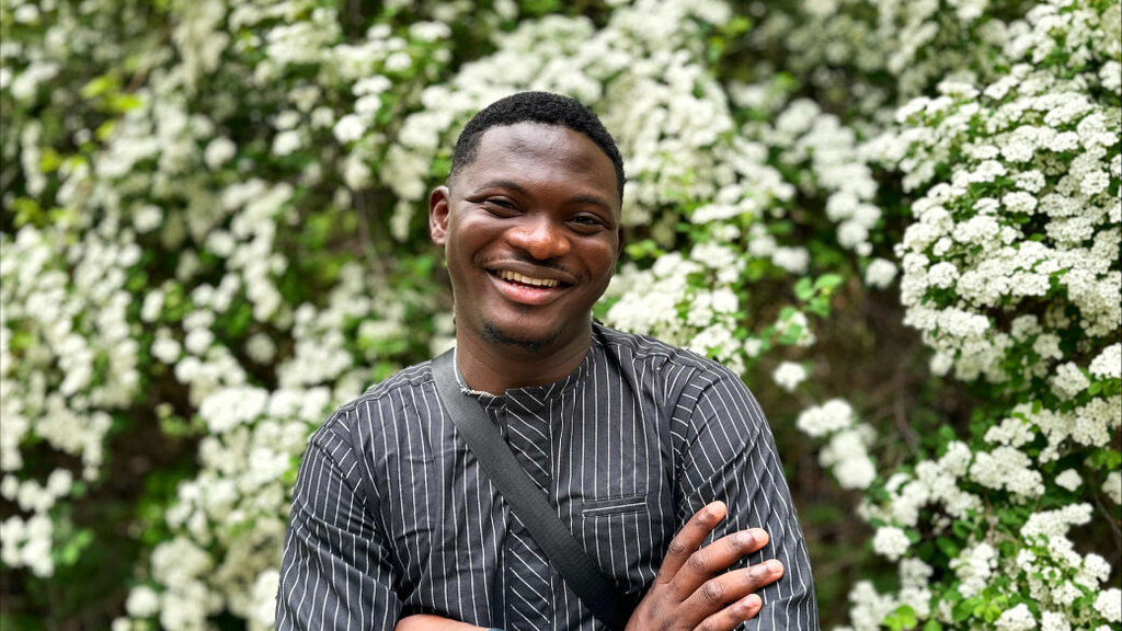 Adedayo Agarau, smiling with his arms crossed in front of a bush covered in white flowers