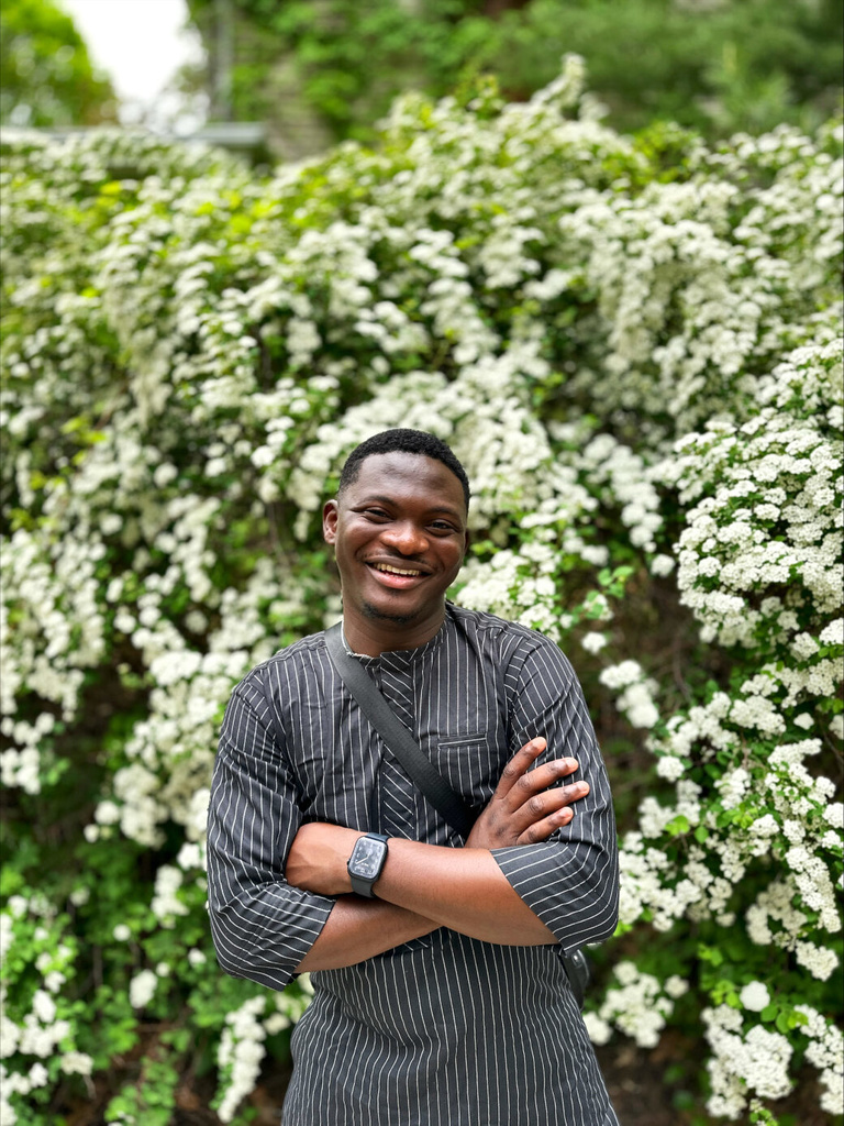 Adedayo Agarau, smiling with his arms crossed in front of a bush covered in white flowers
