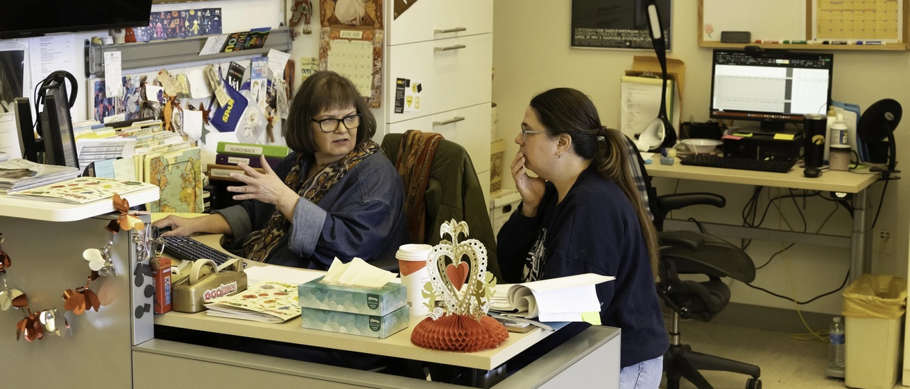 Cindy Kubu sitting at her desk in the costume shop talking to a collaborator