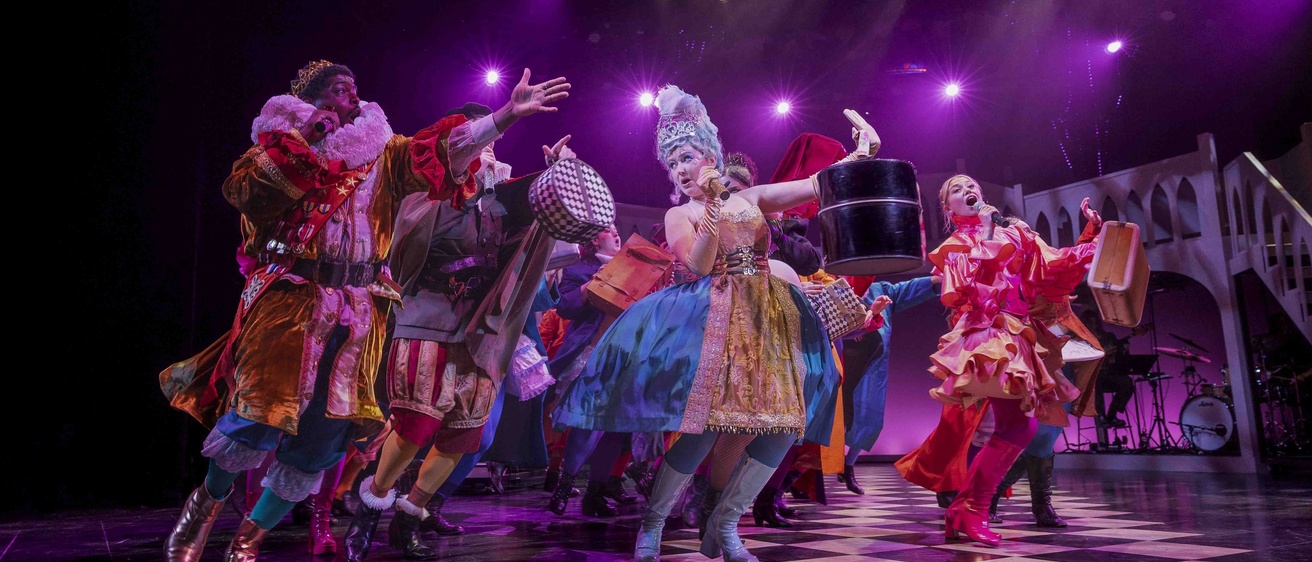 Head Over Heels musical cast members singing into microphones while performing on stage in colorful costumes and dramatic purple lighting