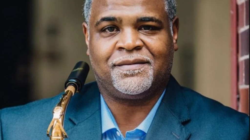 Professor Damani Phillips, wearing a blus shirt and blue suit coat and holding a black and gold saxophone