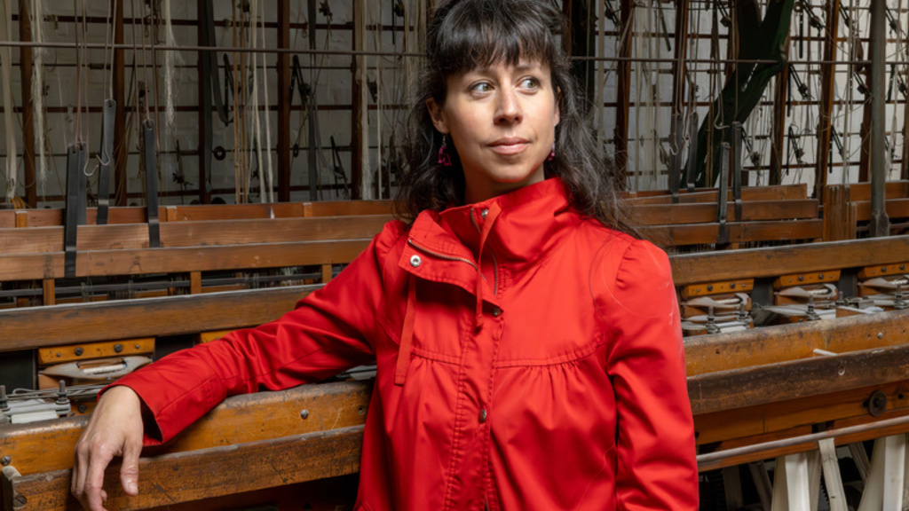 Stephanie Miracle poses in a red jacket with one hand resting on an old wooden pipe organ mechanism, its pipes, wires, and tracker action visible in the background.